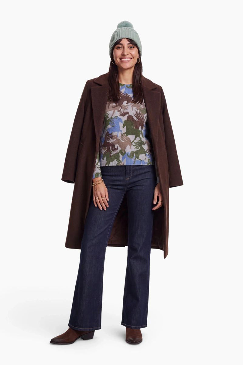 Eine lächelnde Frau trägt eine salbeigrüne, gerippte Strickmütze, einen braunen Wollmantel über einem Pullover mit Pferdemuster und dunkelblaue, ausgestellt geschnittene Jeans. Sie steht vor einem schlichten weißen Hintergrund und rundet ihren Look mit braunen Lederstiefeln ab.