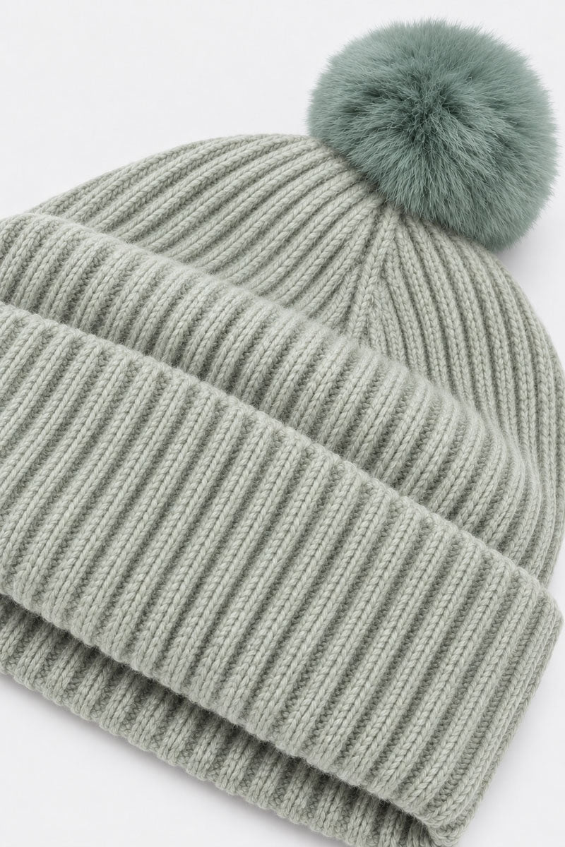 Eine salbeigrüne, gerippte Strickmütze mit einem passenden, flauschigen Bommel. Die Mütze verfügt über einen breiten, umgeschlagenen Rand und besticht durch ihre weiche, strukturierte Optik aus einer Woll-Kaschmir-Mischung vor einem schlichten weißen Hintergrund.