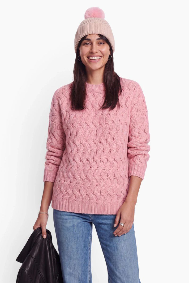 Eine lächelnde Frau trägt einen rosa Zopfmusterpullover, eine hellrosa Rippstrickmütze mit Bommel und eine blaue Jeans. Vor einem schlichten weißen Hintergrund hält sie in einer Hand eine schwarze Jacke.