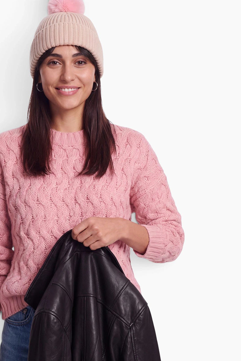 Eine lächelnde Frau trägt einen rosa Rippstrick-Pullover und eine dazu passende Mütze mit Bommel. Vor einem schlichten weißen Hintergrund hält sie eine schwarze Lederjacke vor sich.