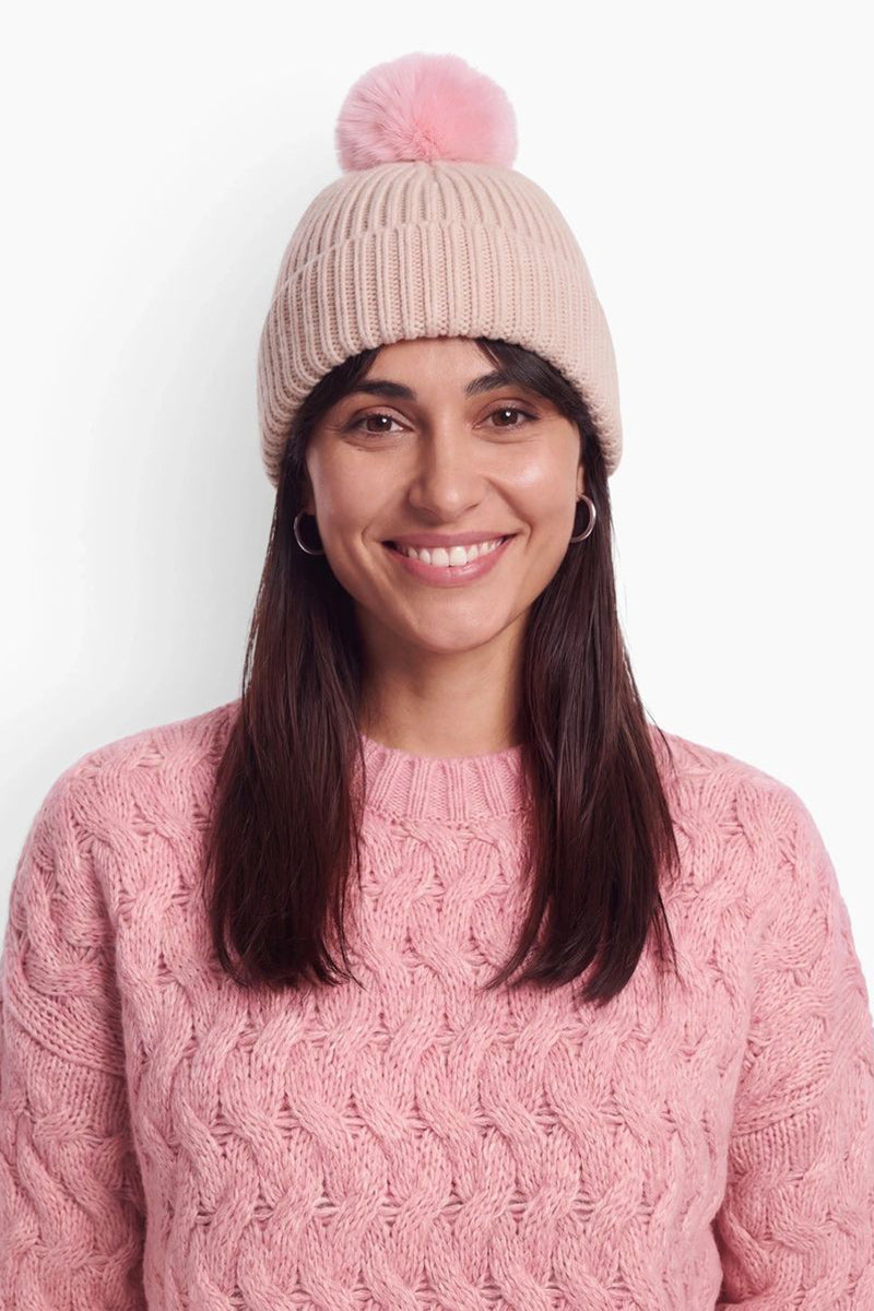 Eine Frau mit dunklem Haar lächelt und trägt eine hellrosa Rippstrick-Mütze mit einem flauschigen Bommel. Vor einem schlichten weißen Hintergrund trägt sie einen dazu passenden rosa Pullover mit Zopfmuster.