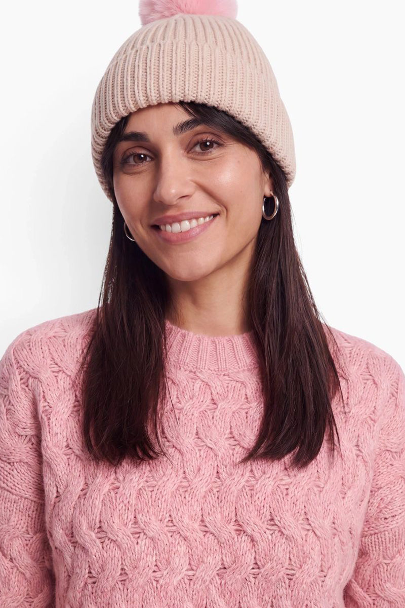 Eine lächelnde Frau mit dunklem Haar trägt eine beige gerippte Mütze mit einem rosa Bommel und einen strukturierten rosa Zopfmusterpullover. Sie trägt kleine silberne Creolen und posiert vor einem schlichten, hellen Hintergrund.