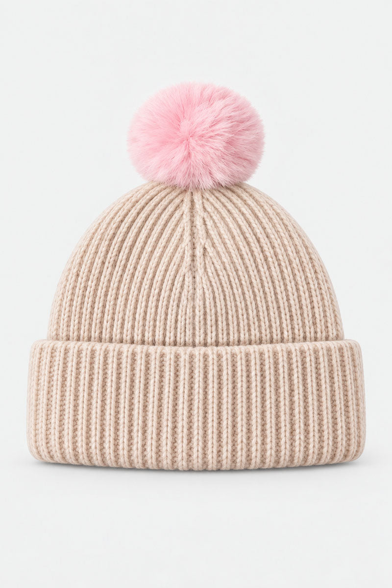 Eine beige Rippstrick-Mütze mit einem breiten, umgeschlagenen Rand und einem großen, flauschigen rosa Bommel oben, mittig auf einem schlichten weißen Hintergrund.