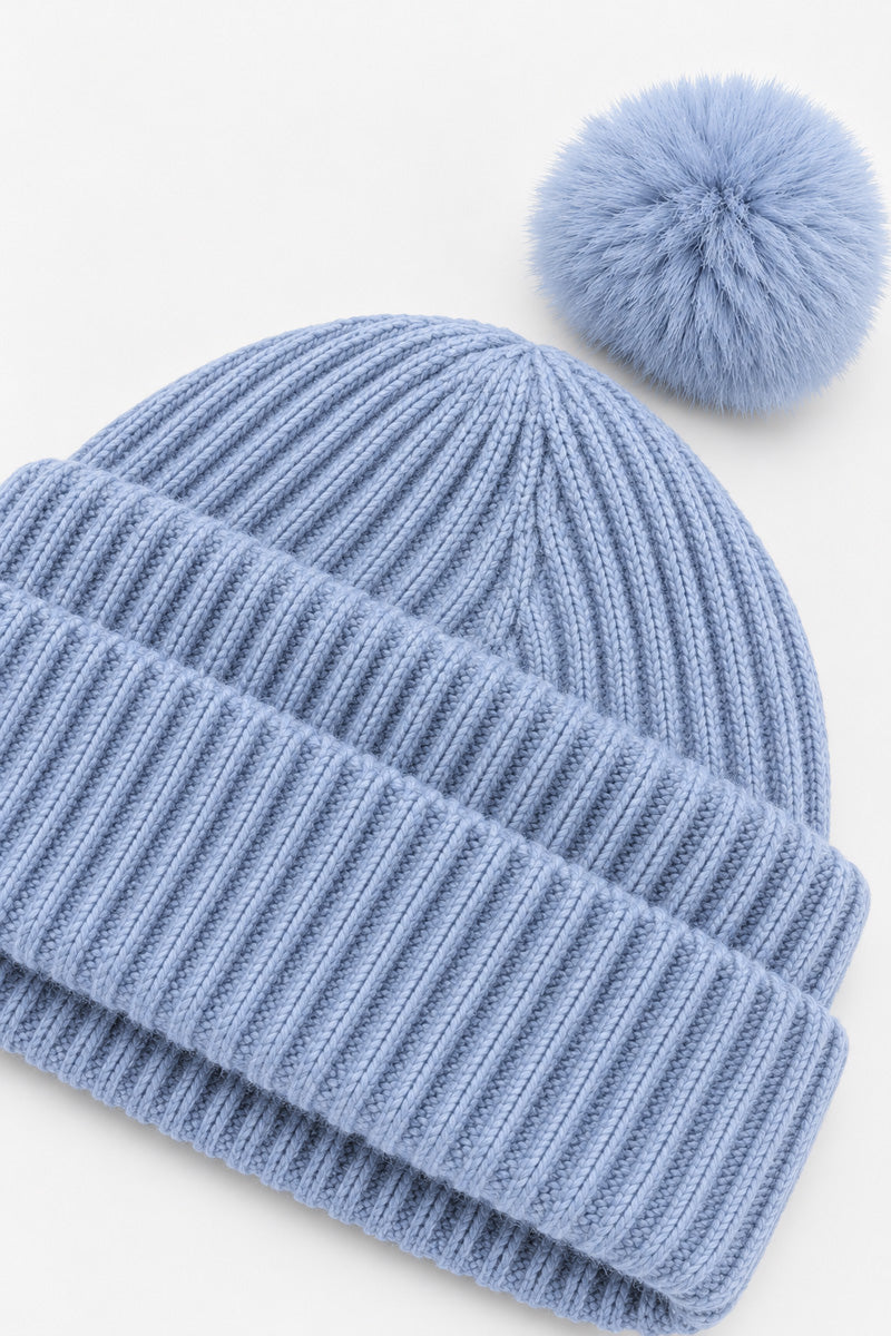 Eine hellblaue Rippstrick-Mütze mit einem passenden Bommel aus Kunstfell oben drauf. Die Wintermütze wird vor einem schlichten weißen Hintergrund gezeigt.