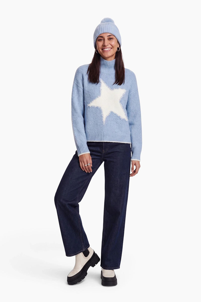 Eine lächelnde Frau trägt einen hellblauen Rollkragenpullover mit einem großen weißen Stern, eine dazu passende gerippte Strickmütze und dunkelblaue Jeans. Abgerundet wird der Look durch weiße Chelsea-Stiefel mit klobigen schwarzen Sohlen vor einem schlichten weißen Hintergrund.