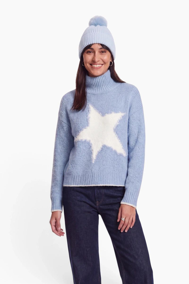 Eine lächelnde Frau trägt einen hellblauen Rollkragenpullover mit einem großen weißen Sternmotiv und eine dazu passende, gerippte Strickmütze mit Bommel. Sie steht vor einem schlichten weißen Hintergrund und trägt eine dunkle Jeans.