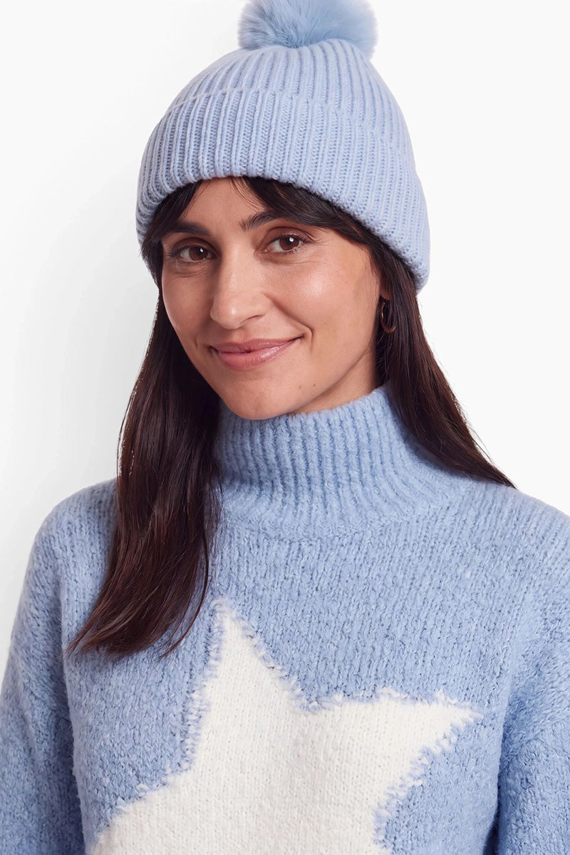 Eine Frau mit dunklem Haar lächelt und trägt eine hellblaue, gerippte Mütze mit Bommel. Sie trägt einen dazu passenden hellblauen Rollkragenpullover mit einem großen, flauschigen weißen Sternenmuster vor einem schlichten weißen Studiohintergrund.