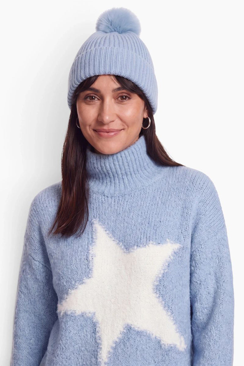 Eine lächelnde Frau mit dunklem Haar trägt eine hellblaue, gerippte Strickmütze mit einem passenden Bommel und einen kuscheligen Rollkragenpullover, auf dessen Vorderseite ein großer, flauschiger weißer Stern vor einem schlichten, hellen Hintergrund zu sehen ist.