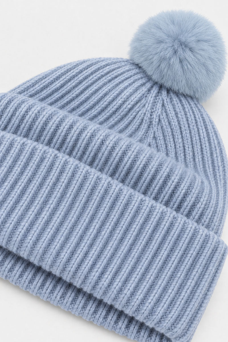 Eine hellblaue, gerippte Strickmütze aus Wolle und Kaschmir mit einem passenden, flauschigen Bommel oben drauf. Die Wintermütze hat einen dicken, umgeschlagenen Rand und eine weiche, strukturierte Optik.