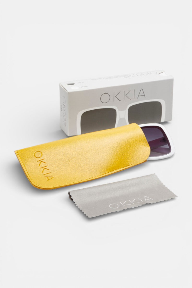 Okkia White Oversize quadratische Sonnenbrille mit dunklen Gläsern (UV 400 Schutz), präsentiert neben einem gelben Okkia-Etui, grauem Reinigungstuch und illustrierter weißer Okkia-Box auf weißem Hintergrund.
