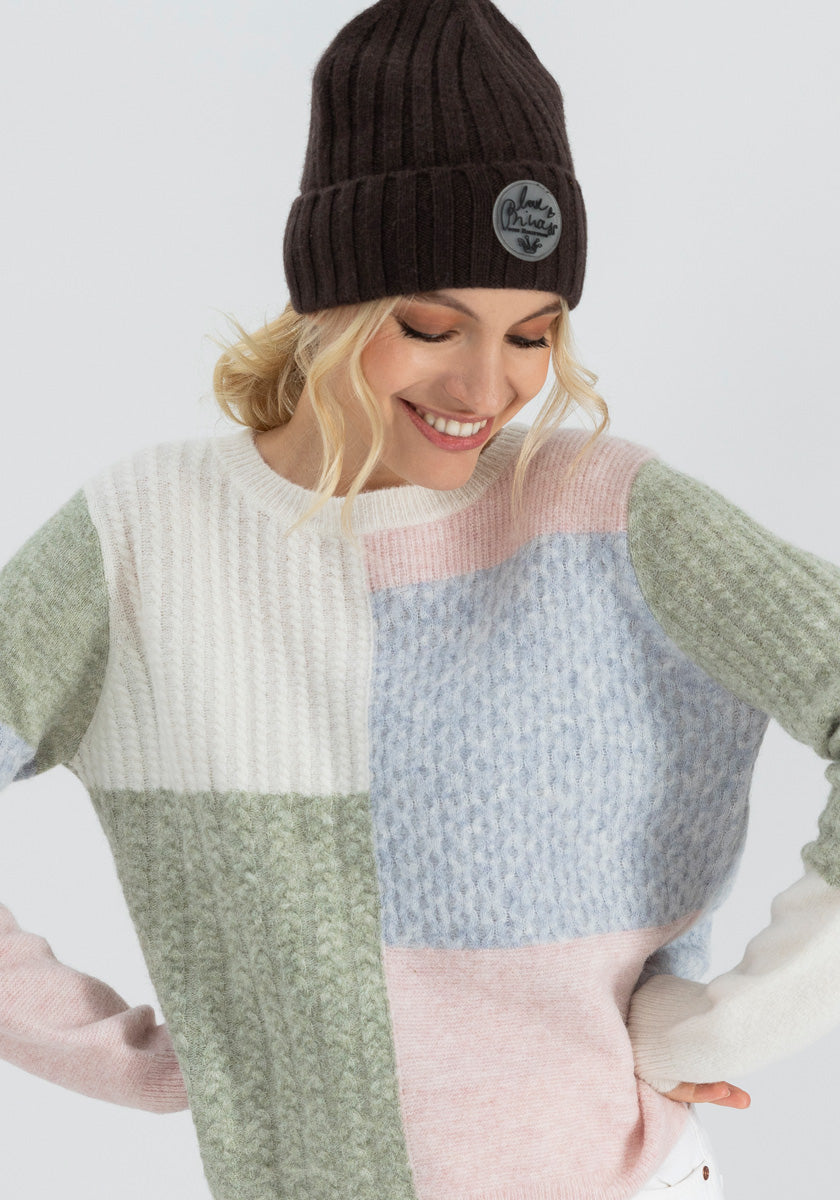 A smiling woman wears a geripptes Design dark brown knit beanie and a textured color-block sweater with Farbiges Strickdetail in white, green, blue, and pink from Princess Goes Hollywood. Her blonde hair is tucked under the hat against a plain light background.