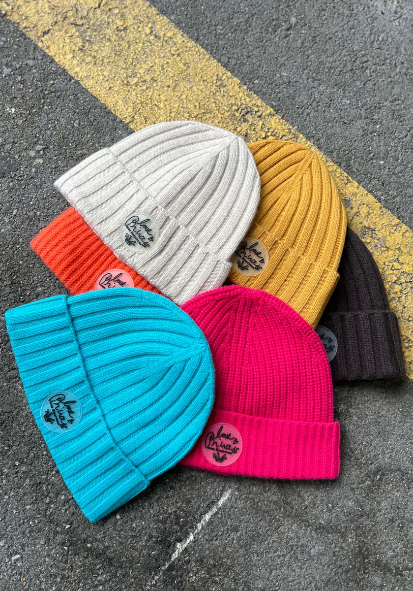 Five ribbed knit beanies by Princess Goes Hollywood in beige, mustard yellow, brown, blue, and magenta with colorful knit detail are fanned out on gray pavement. Each has a round patch with black script and a leaf graphic.