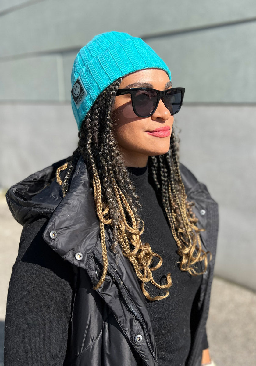 A woman stands outdoors in sunlight, wearing a bright turquoise Princess Goes Hollywood knit beanie with colorful knit detail, black sunglasses, a black puffer vest, and a black top. Long blonde-tipped braids drape over her shoulders as she smiles slightly.