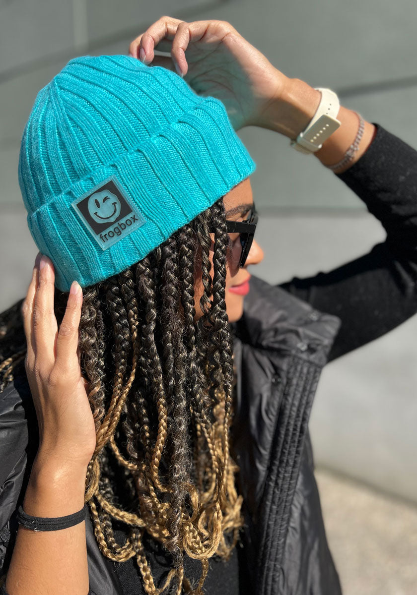 A person with long, braided hair wears a bright turquoise beanie with colorful knit detail from Princess Goes Hollywood, black sunglasses, a white smartwatch, and a black puffy vest, posing against a light gray background in sunlight.