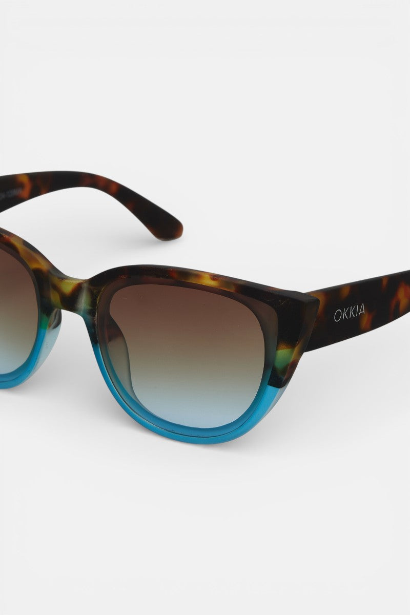 Die Cat-Eye-Sonnenbrille von Okkia hat einen schildpattfarbenen oberen Rahmen, der in ein durchscheinendes Blau übergeht, UV-Schutzgläser mit Farbverlauf und das Okkia-Logo in Weiß auf dem linken Bügel.