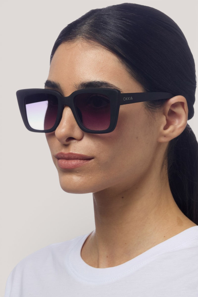 Eine Frau mit langen dunklen Haaren trägt eine schwarze Okkia ELBA Sonnenbrille in Übergröße mit UV-400-Schutz und Verlaufsgläsern, gepaart mit einem weißen T-Shirt. An der Seite steht "Okkia", während sie vor einem schlichten hellen Hintergrund posiert und nach links schaut.
