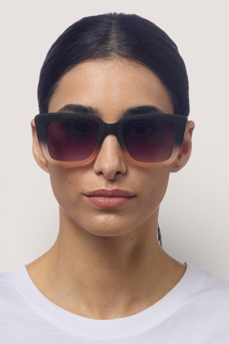 Eine Frau mit glattem, dunklem Haar trägt eine schwarze Okkia Oversize-Design-Sonnenbrille mit Verlaufsgläsern und ein weißes Hemd und blickt mit neutralem Gesichtsausdruck in die Kamera vor einem schlichten, hellen Hintergrund.