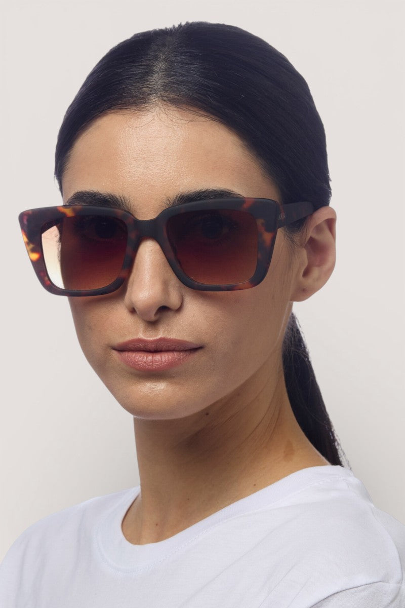 Eine Frau mit glattem, schwarzem Haar trägt eine Okkia Oversize Sonnenbrille mit UV 400 Schutz und Soft-Touch Gestell, gepaart mit einem weißen Rundhalsshirt. Sie blickt mit neutralem Gesichtsausdruck vor einem schlichten, hellen Hintergrund nach vorne.