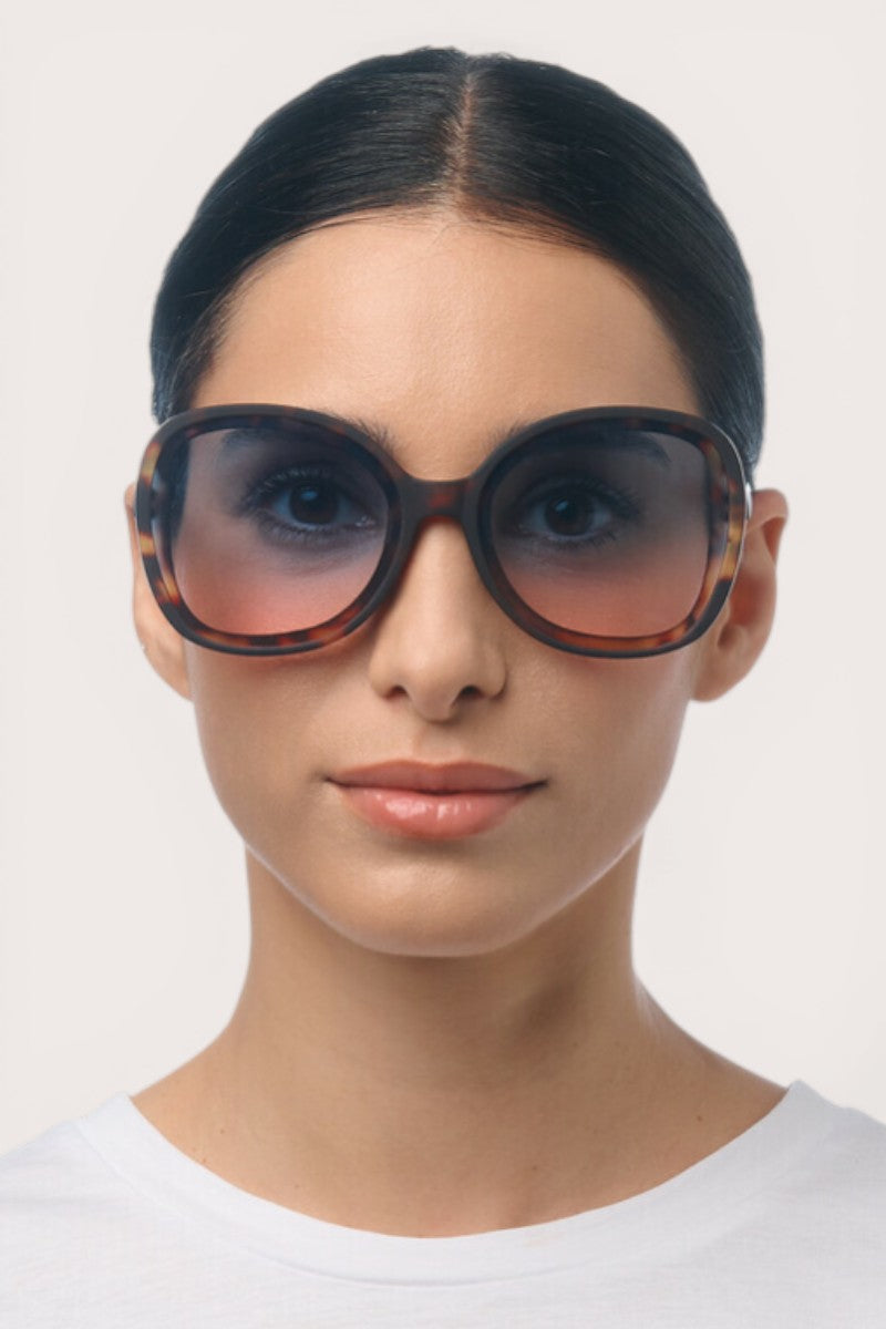 Eine Frau mit dunklem, nach hinten gegeltem Haar trägt eine übergroße, braune Okkia-Sonnenbrille mit UV-400-Schutz und ein weißes Hemd. Sie blickt mit neutralem Gesichtsausdruck vor einem schlichten, hellen Hintergrund nach vorn.