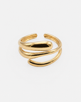 Ein glänzender goldener Ring von Princess Goes Hollywood mit einem modernen, offenen Design aus drei geschwungenen Bändern, die sich überlappen und umeinander winden und so einen eleganten, minimalistischen Look ergeben. Dieses moderne Schmuckstück aus hochwertigem Edelstahl ist zudem ein verstellbarer Ring, der sich perfekt anpassen lässt. Der Hintergrund ist schlicht weiß, wodurch die glatte Metalloberfläche des Rings besonders gut zur Geltung kommt.