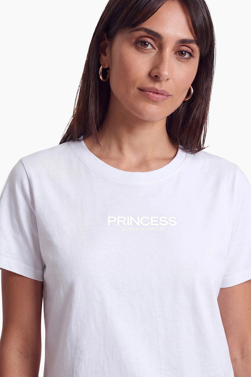 Eine Frau mit schulterlangem braunem Haar trägt ein schlichtes weißes Baumwoll-T-Shirt mit Rundhalsausschnitt der Marke Princess Goes Hollywood. Der Markenname ist in weißer Schrift auf ihrer Brust aufgedruckt, und sie rundet den Look mit kleinen goldenen Creolen ab.