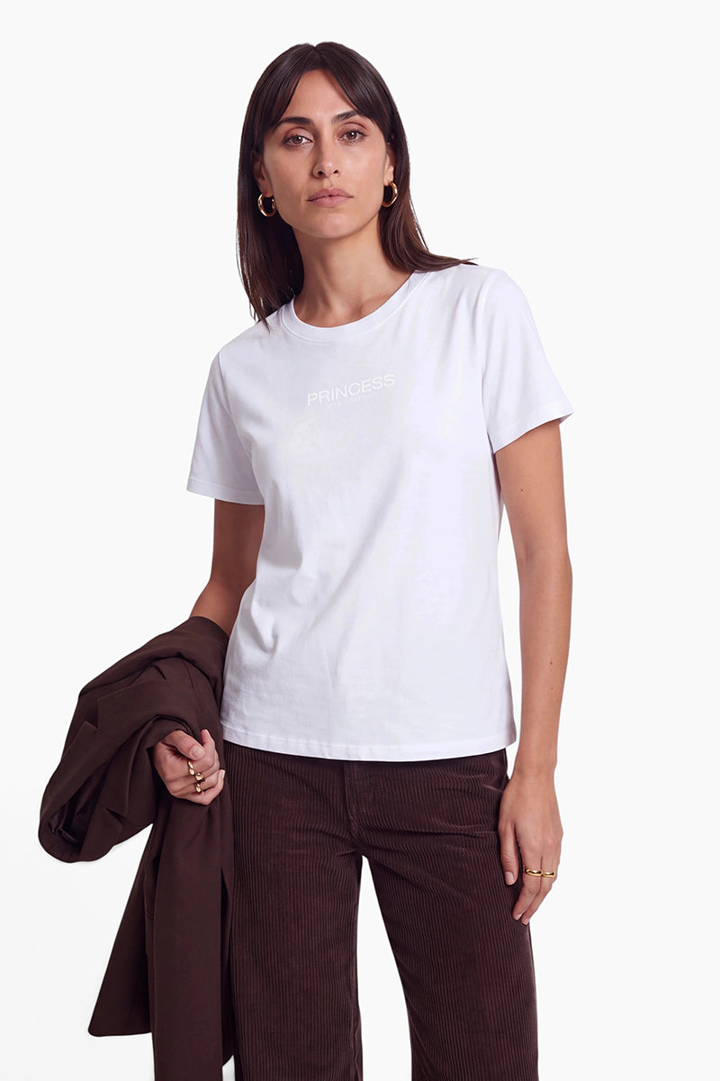 Vor einem weißen Hintergrund steht eine Frau mit dunklem Haar und goldenen Creolen, die ein Baumwoll-T-Shirt mit der Aufschrift Princess Goes Hollywood trägt. Sie hält eine dunkelbraune Jacke in der Hand und trägt dazu eine passende dunkelbraune Cordhose.