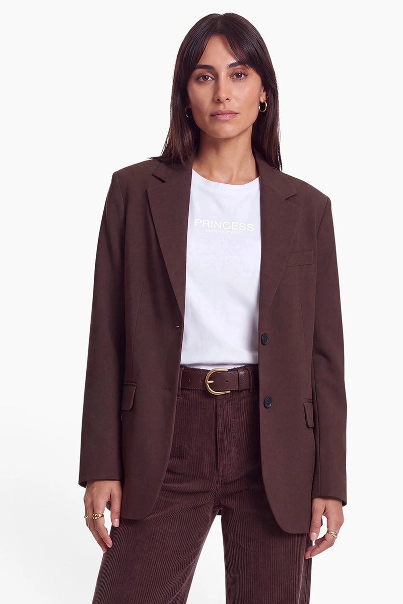 Eine Frau mit langen braunen Haaren posiert in einem schokoladenbraunen Blazer über einem weißen Princess Goes Hollywood-T-Shirt. Sie trägt einen passenden braunen Gürtel und eine Cordhose vor einem schlichten weißen Hintergrund.