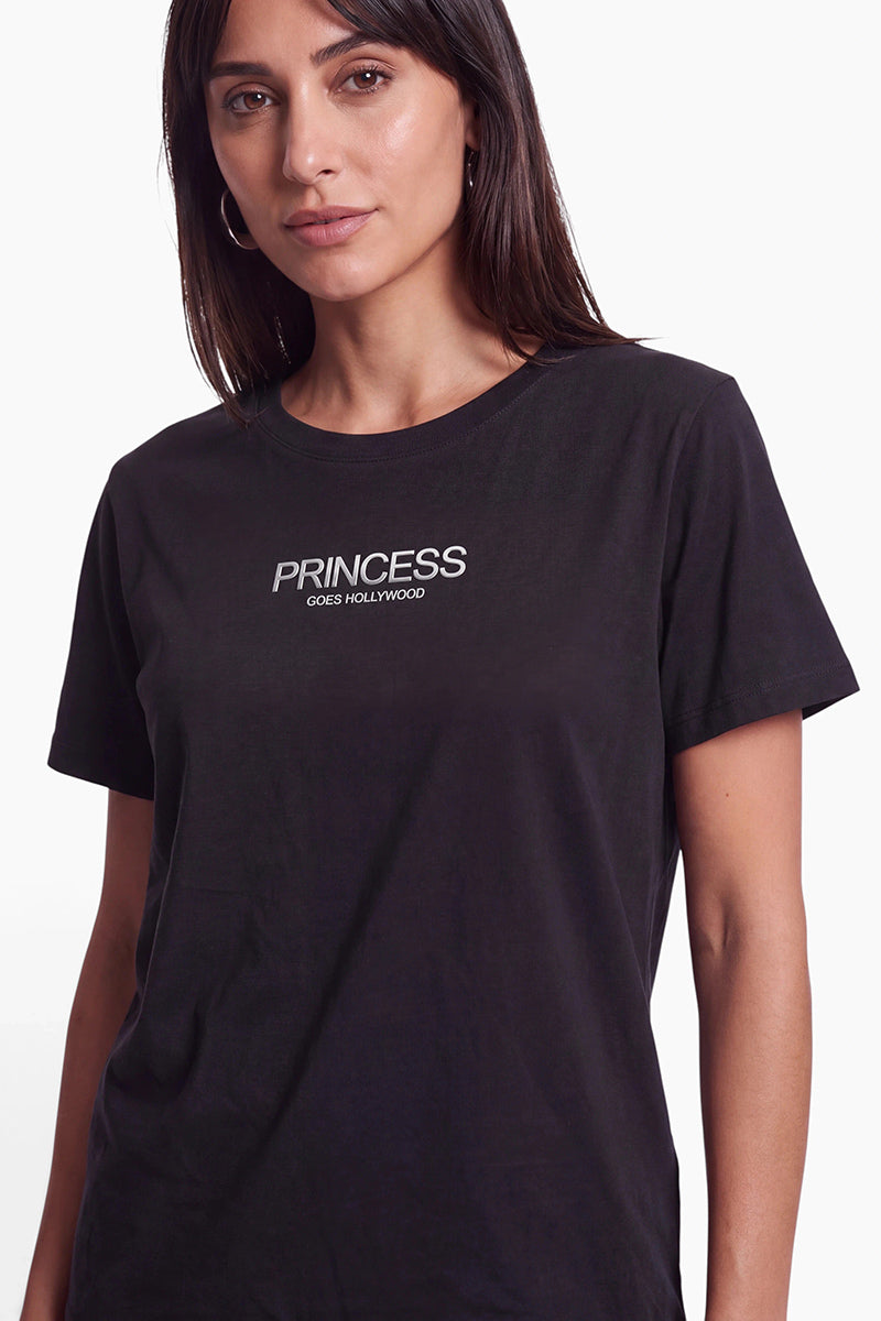 Eine Frau mit dunklem Haar trägt ein schwarzes Baumwoll-T-Shirt, auf dessen Brust der weiße Schriftzug PRINCESS GOES HOLLYWOOD aufgedruckt ist. Sie hat einen neutralen Gesichtsausdruck und trägt silberne Creolen vor einem schlichten weißen Hintergrund.