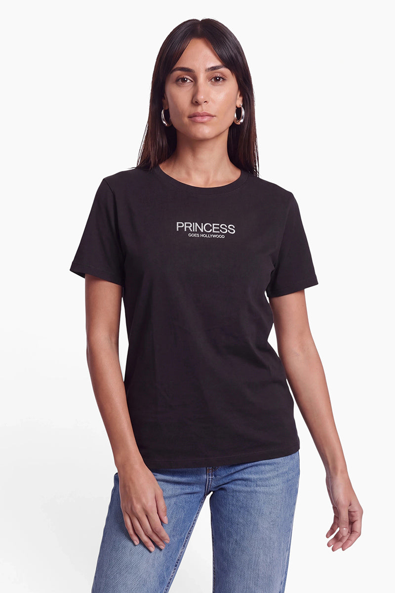 Eine Frau mit langen, dunklen Haaren trägt ein schwarzes Princess Goes Hollywood-T-Shirt mit weißer Aufschrift. Sie hat silberne Creolen und trägt eine blaue Jeans vor einem schlichten weißen Hintergrund.