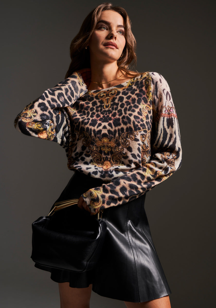 A woman poses confidently before a dark background in a Princess Goes Hollywood animal- and baroque-print long-sleeve top, a high-waisted black leather mini skirt, and a black handbag, her gaze to the side and one hand in her hair.