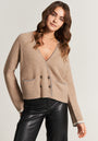 A woman with long wavy brown hair wears a beige Princess Goes Hollywood wool-cashmere cardigan with double-breasted buttons and glitter details on the pockets and cuffs, paired with black leather pants and a gold chain necklace against a plain background.
