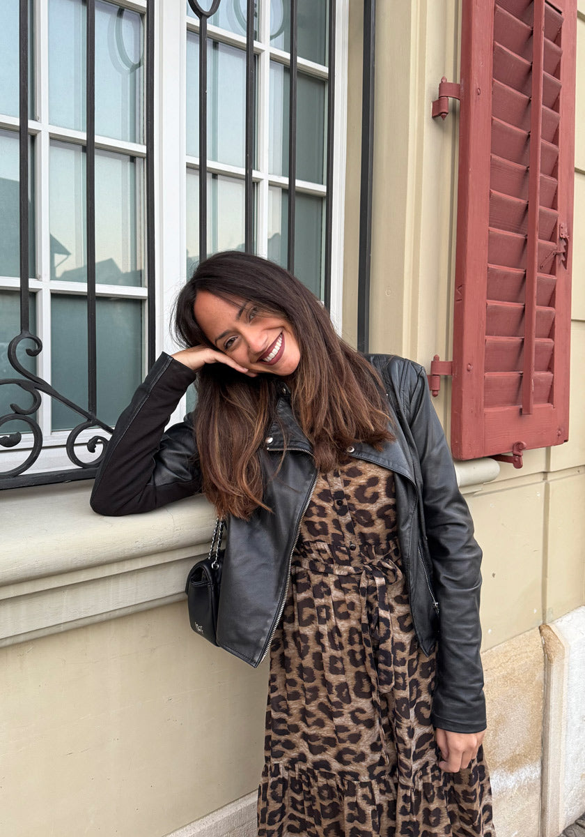 A woman with long brown hair wears a black leather jacket and a Princess Goes Hollywood leopard-print midi dress. Smiling, she leans her head on her hand in front of a cream-colored building with black-barred windows and red shutters.
