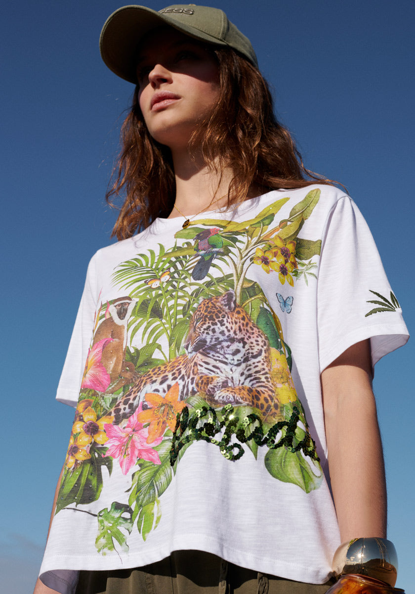A woman stands outdoors against a blue sky, wearing a green cap and the Princess Goes Hollywood T-Shirt mit Tropical Print with Paradise in green strass on white, plus wavy hair and a gold bracelet.