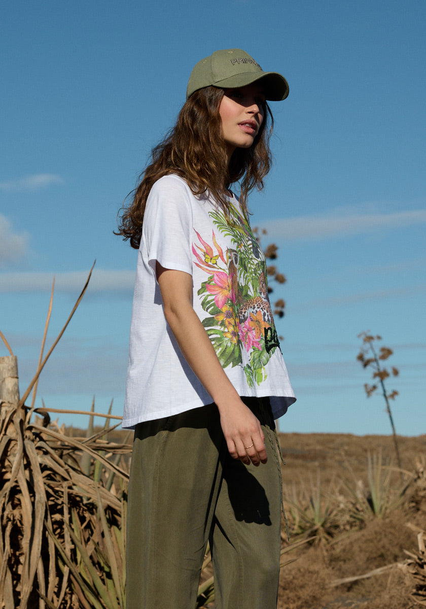 A woman stands outdoors in sunlight, wearing olive-green pants and a Princess Goes Hollywood T-Shirt mit Tropical Print. Dry plants and a blue sky with wispy clouds are in the background as she looks to the side, partially in shadow.