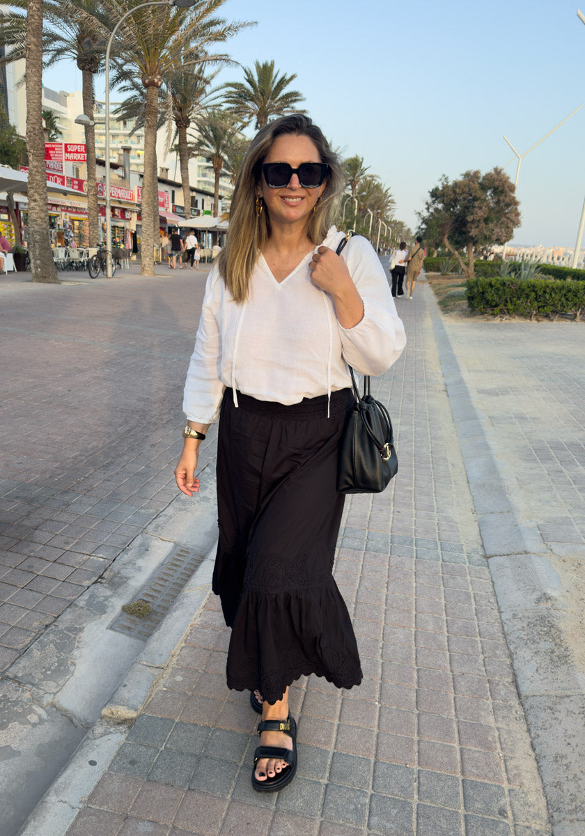 A smiling woman walks on a palm-lined promenade wearing a white Princess Goes Hollywood Leinen-Bluse with Rüschen, black wide-leg pants, black sandals, sunglasses, and a black shoulder bag. Shops and people are in the sunny background.