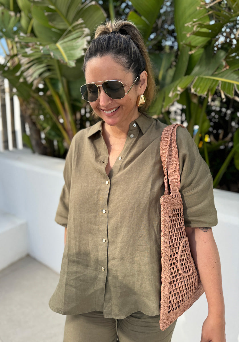 A woman with a ponytail smiles outdoors, wearing aviator sunglasses and a loose Princess Goes Hollywood Leinen-Hemdbluse mit 3/4-Ärmel with pants. She carries a pink woven tote. Large green tropical leaves and white walls are in the background.