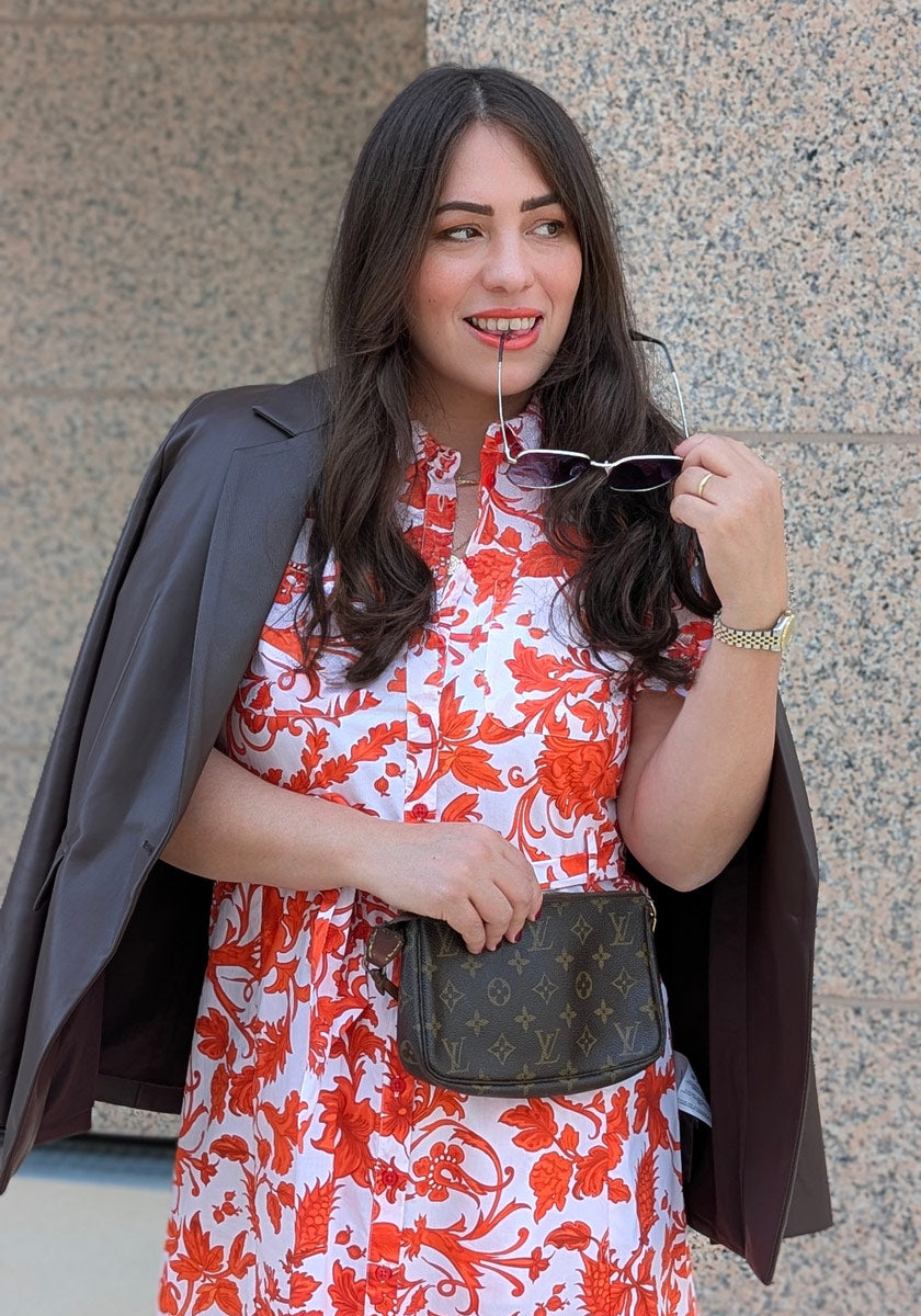 A woman with long dark hair in a Princess Goes Hollywood floral maxi dress holds sunglasses near her mouth, smiling. She layers a dark blazer over her shoulders, wears a gold watch, carries a small Louis Vuitton purse, and stands by a stone wall.