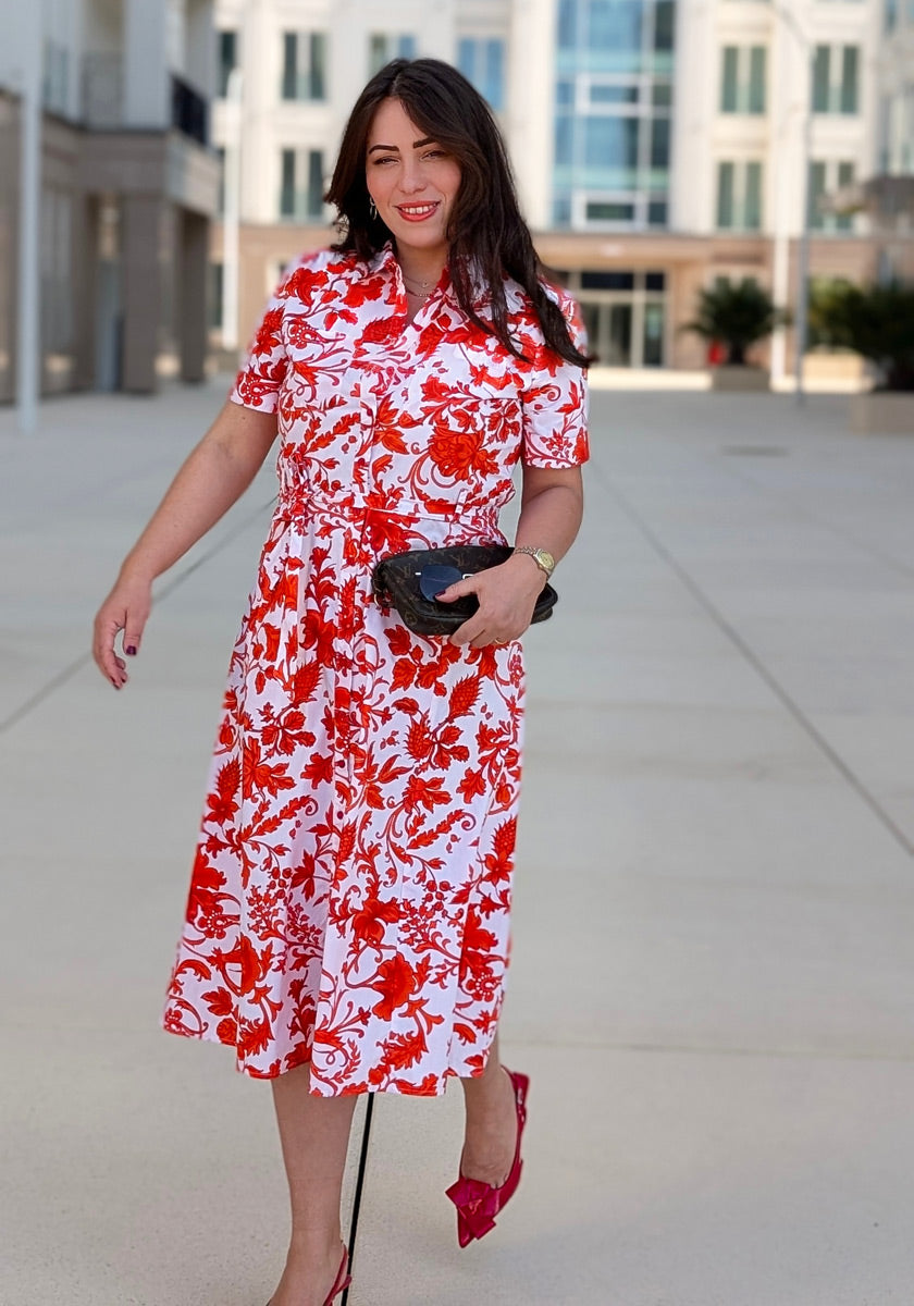 A woman with long brown hair wears a white Princess Goes Hollywood maxi dress with red floral prints, red pointed heels, and holds a black clutch. She smiles outdoors on a sunny day with modern buildings in the background.
