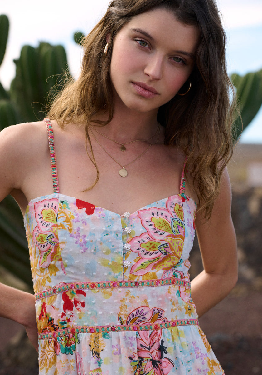 A young woman stands outdoors before cacti, wearing the Florales Trägerkleid aus Baumwolle by Princess Goes Hollywood. She has wavy brown hair, gold hoop earrings, layered necklaces, and gazes neutrally at the camera in soft light.