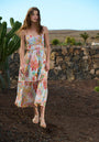 A woman with long, wavy brown hair stands outdoors on rocky ground wearing the Florales Trägerkleid aus Baumwolle by Princess Goes Hollywood. She pairs it with sandals; a stone wall, cactus, and clear sky complete the scene.