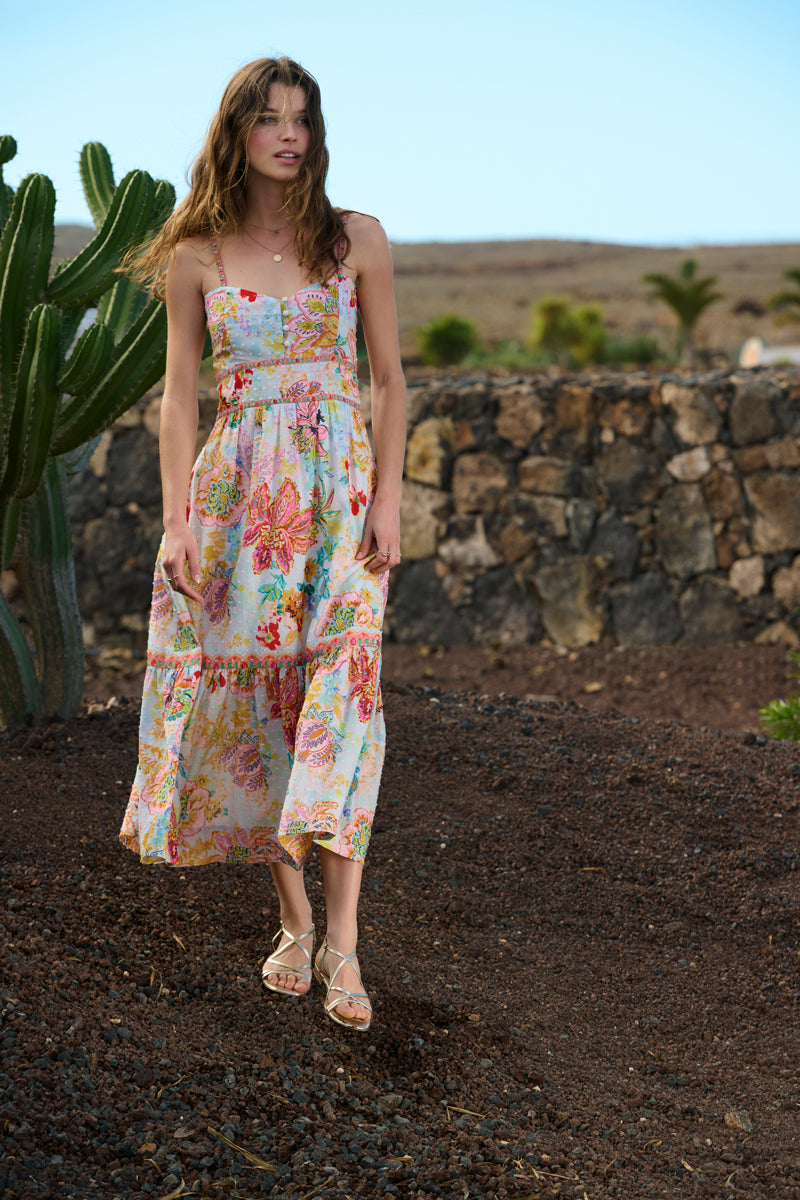 A woman in a colorful, floral sundress and sandals stands outdoors on rocky ground near a stone wall and large cactus. Her brown hair is loose, and she looks toward the camera, with a dry landscape and blue sky in the background.