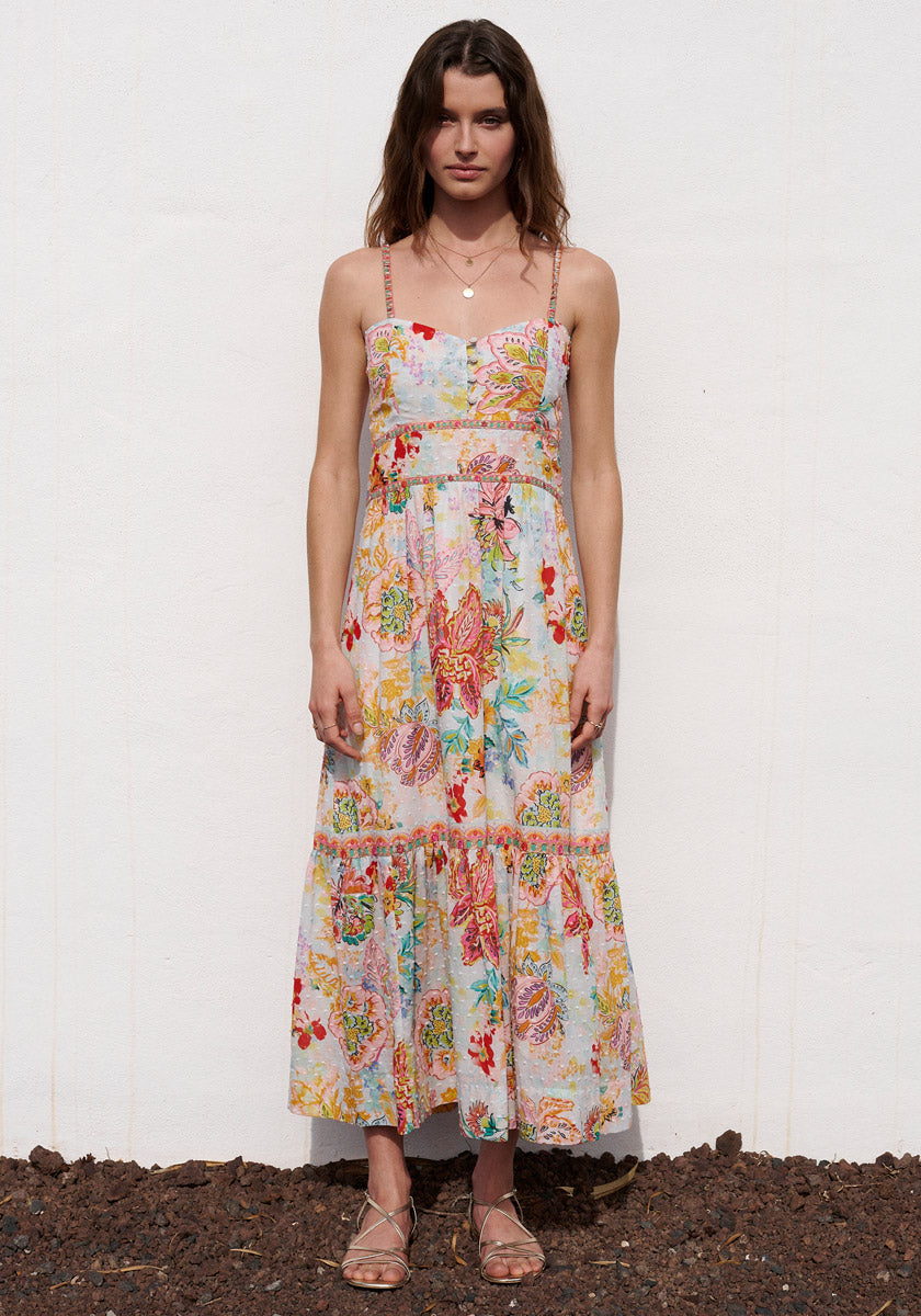 A woman with wavy brown hair stands on dark stones against a white wall, wearing the Florales Trägerkleid aus Baumwolle by Princess Goes Hollywood with dünnen Trägern, Stufenrock, beige Sandalen und goldener Kette.