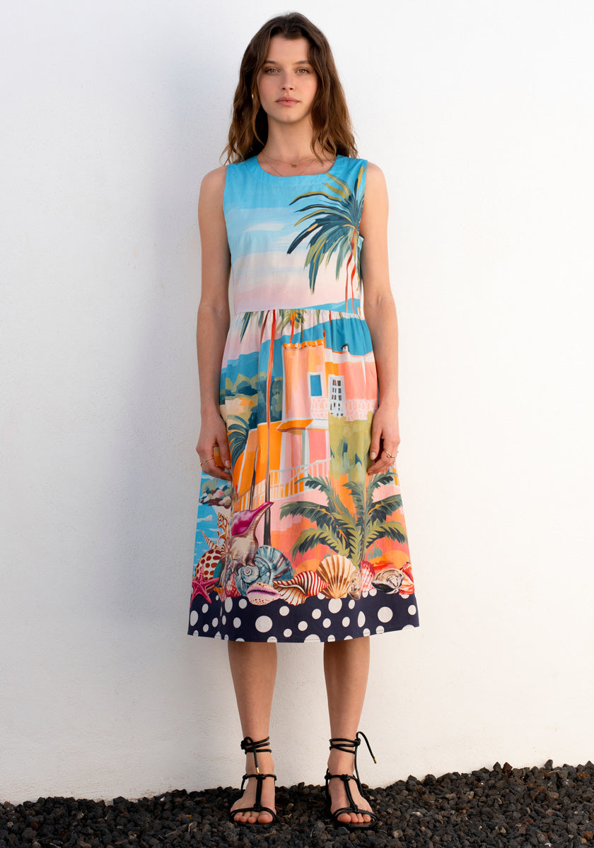 A woman in the Ärmelloses Kleid Miami by Princess Goes Hollywood, featuring a vivid Miami print and a navy-blue polka-dot hem, stands against a white wall with loose brown hair and black strappy sandals.