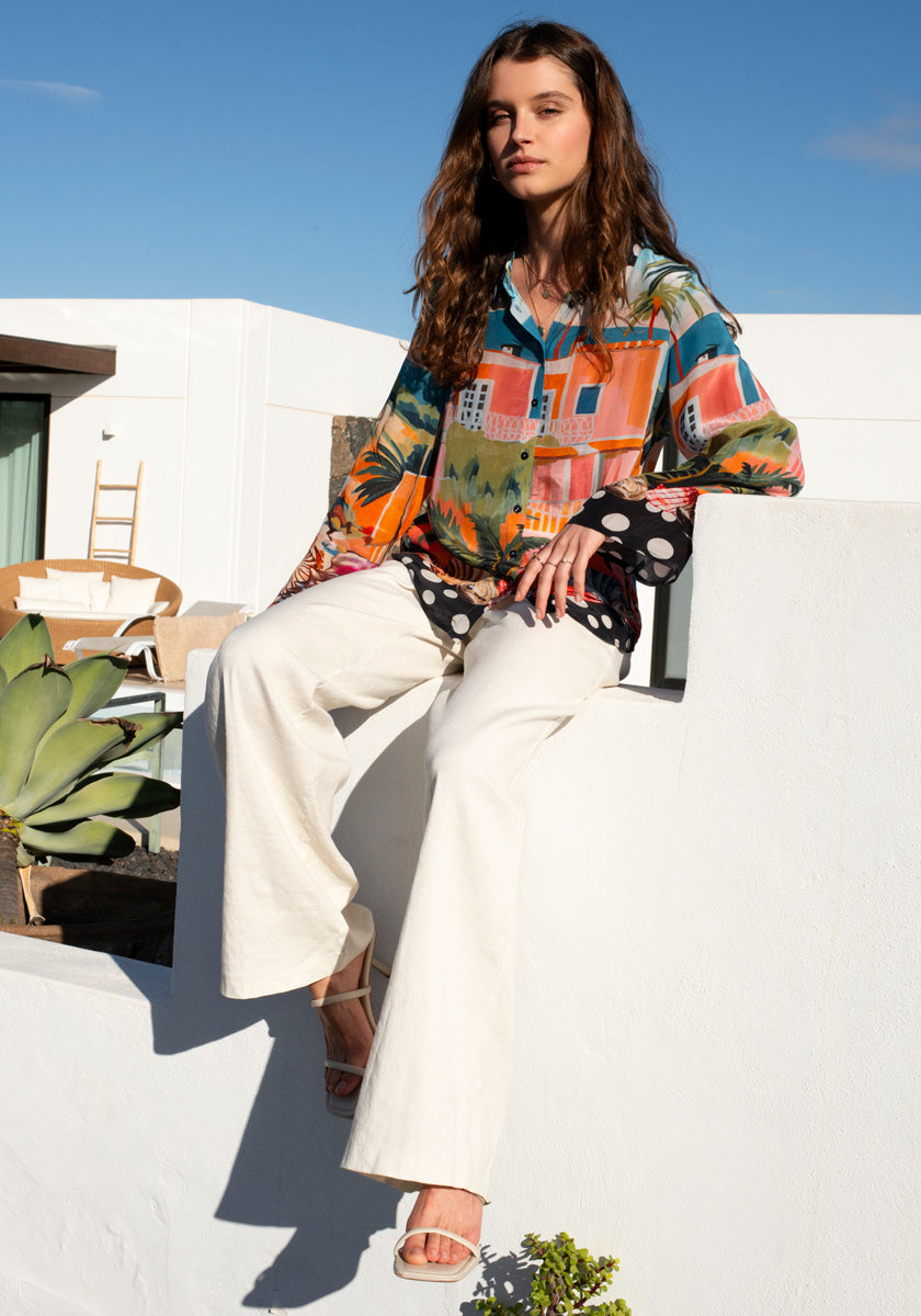 A woman with long brown hair sits on a white wall outdoors, wearing the Bluse Miami by Princess Goes Hollywood, wide-leg white pants, and white sandals. A modern building, wooden chair, and succulents complete the sunny scene.