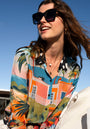 A smiling woman with wavy brown hair wears the Princess Goes Hollywood Bluse Miami with vibrant print and langen weiten Ärmeln, accessorized with large black sunglasses and layered gold necklaces, outdoors under a clear blue sky.