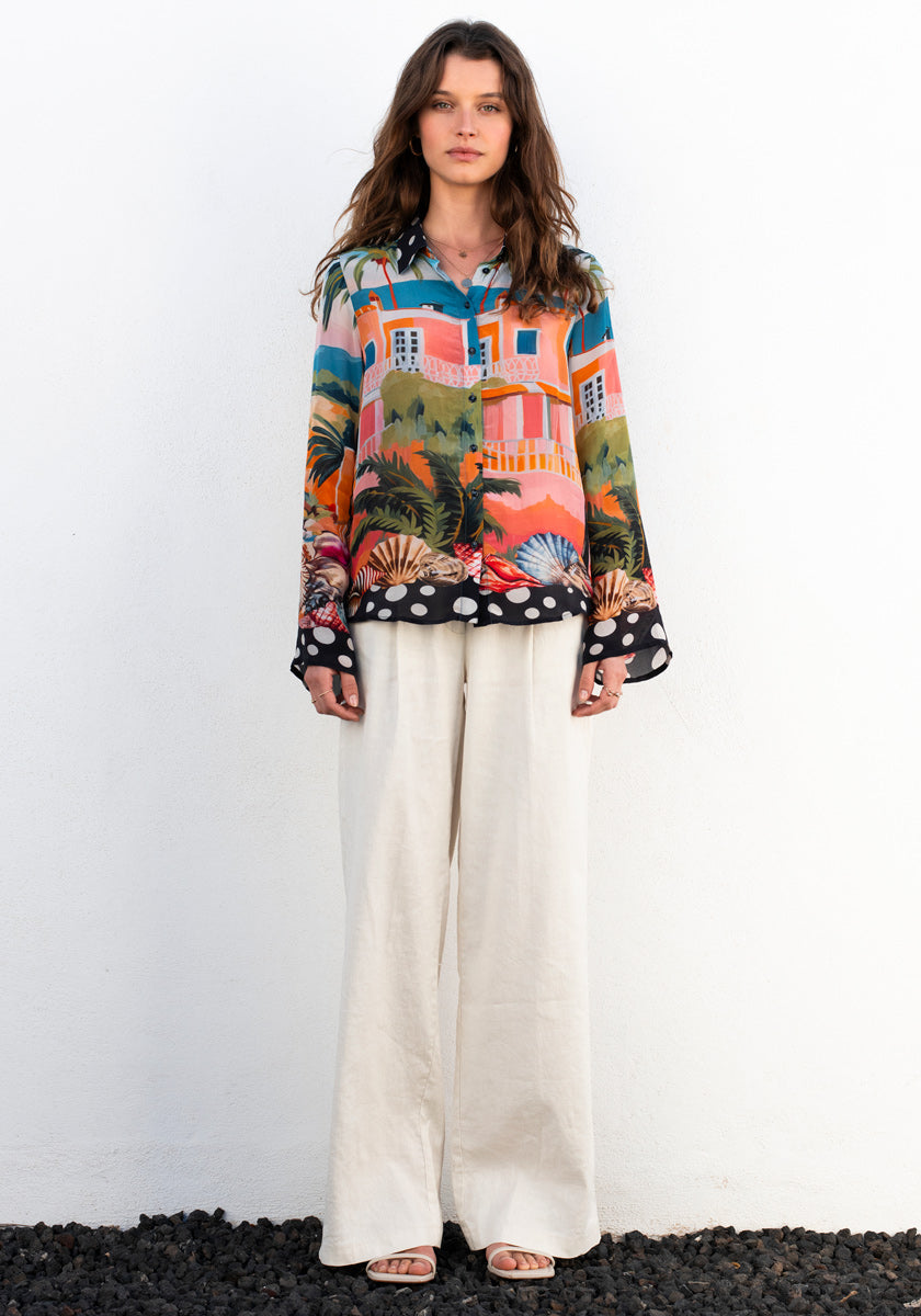 A woman stands against a white wall, wearing the Princess Goes Hollywood Bluse Miami with tropical house and palm tree print, featuring long wide sleeves. She pairs it with loose cream wide-leg pants and white sandals, standing on black pebbles.