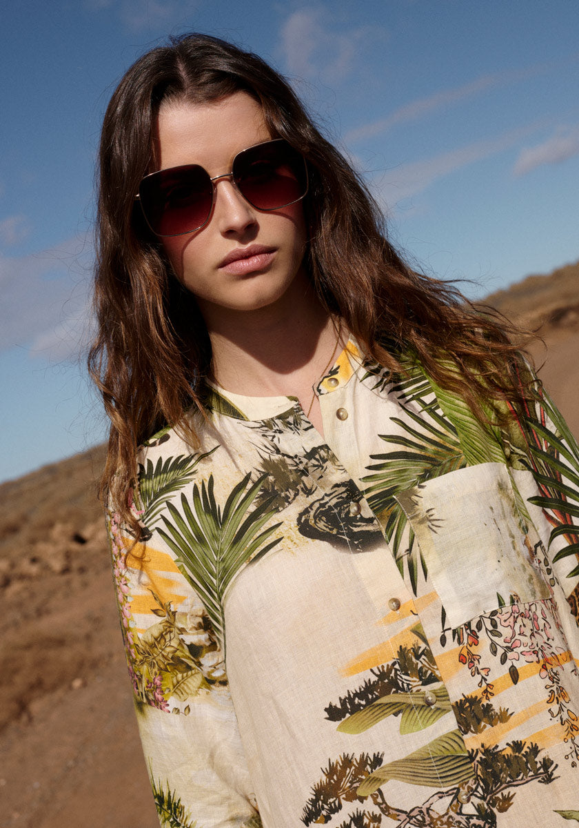A young woman with long brown hair and sunglasses stands outdoors under a blue sky, wearing the Princess Goes Hollywood Leinenkleid Safari. The dry, rocky landscape and distant hills provide a striking backdrop.