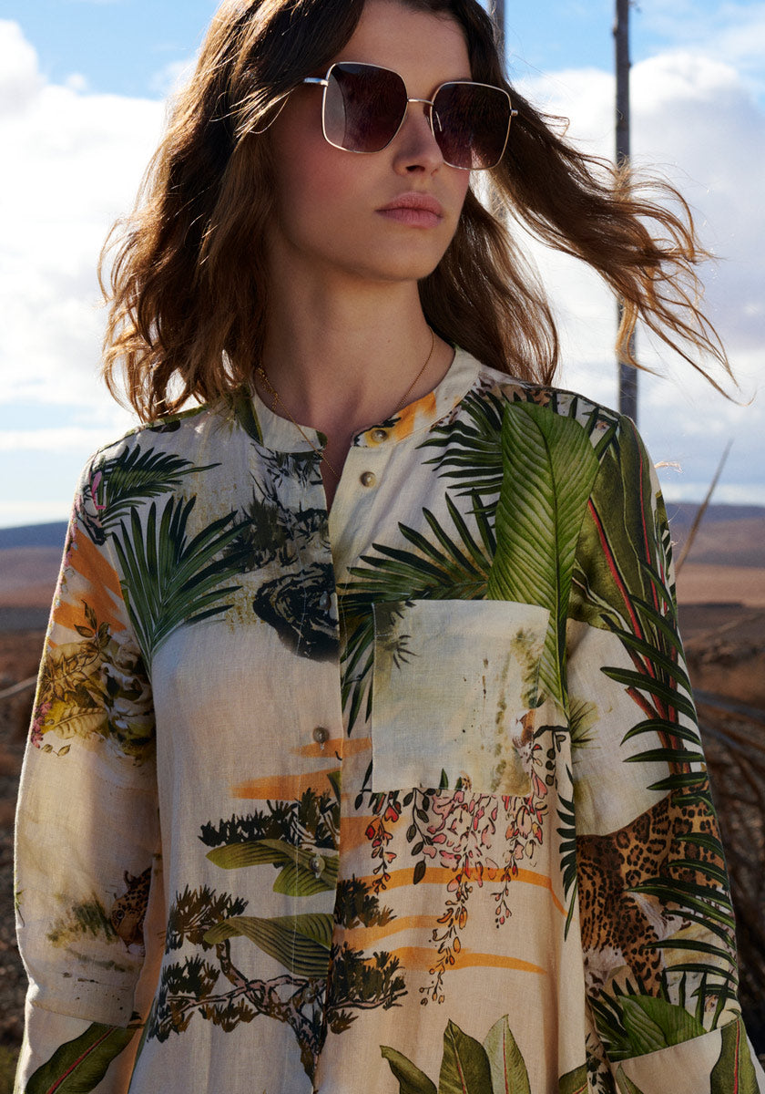 A woman with wavy brown hair wears large square sunglasses and the cream Leinenkleid Safari by Princess Goes Hollywood, featuring green palm leaf, leopard, and floral safari print. She stands outdoors with sunlight on her face and sand behind her.