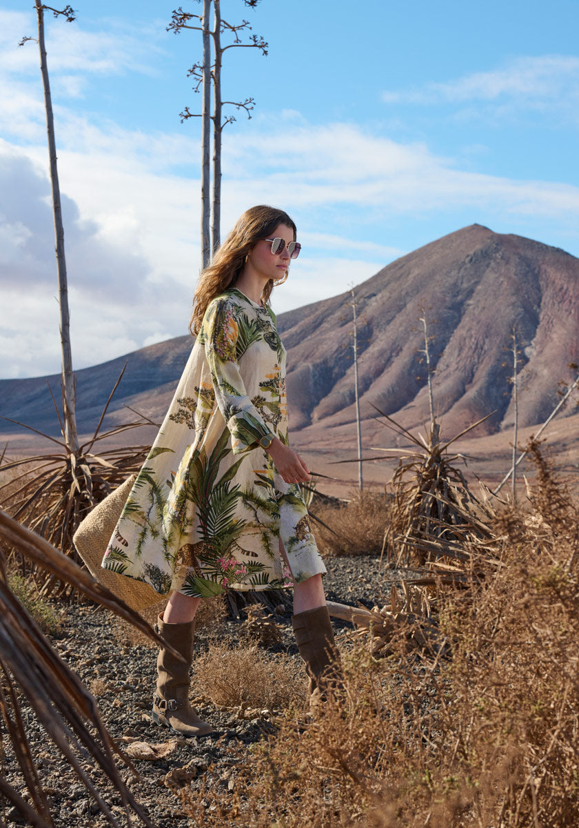 A woman in the Leinenkleid Safari by Princess Goes Hollywood and tall boots walks on a rocky, dry mountain path with sparse plants under a partly cloudy sky, wearing sunglasses as she looks ahead.