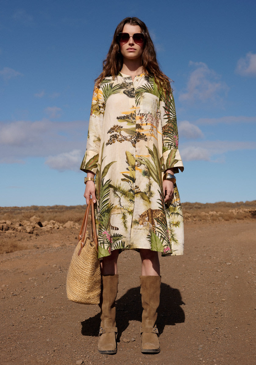 A woman stands on a dirt path under a blue sky, wearing the knee-length Leinenkleid Safari by Princess Goes Hollywood, brown knee-high boots, sunglasses, and carrying a woven straw tote bag across the dry, rocky terrain with scattered clouds.
